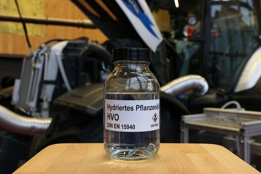 Eine Laborflasche mit transparenter Flüssigkeit steht vor einem Traktor mit geöffneter Motorhaube.